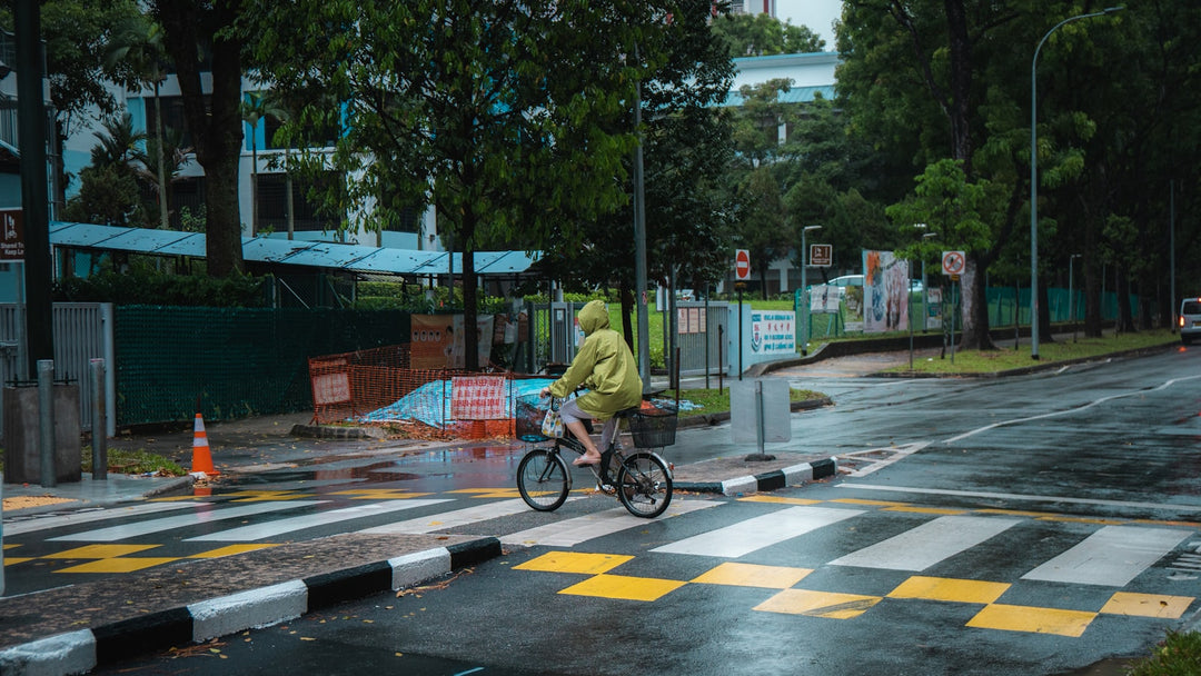 Fietser in felgele regenjas steekt een natte weg over, van veraf zichtbaar bij daglicht